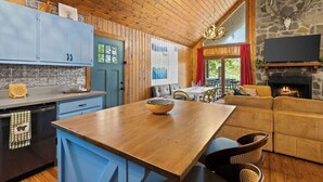 A cozy cabin interior with a wooden ceiling, stone fireplace, and a dining area with a large table and sofa.
