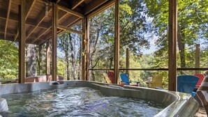 A hot tub in a wooden structure with large windows, surrounded by trees and outdoor chairs.