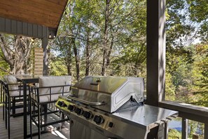 En uteplats med en grill i rostfritt stål, bekväma sittplatser och utsikt över ett skogsområde.