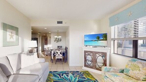 Una sala de estar con sofá, un sofá estampado con motivos florales, un televisor sobre un mueble bajo y una zona de comedor con sillas y una mesa.