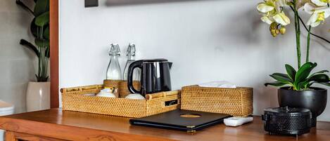 A wooden desk with a black coffee maker, a glass bottle, a woven basket, a black mug, a white remote, and a potted orchid.