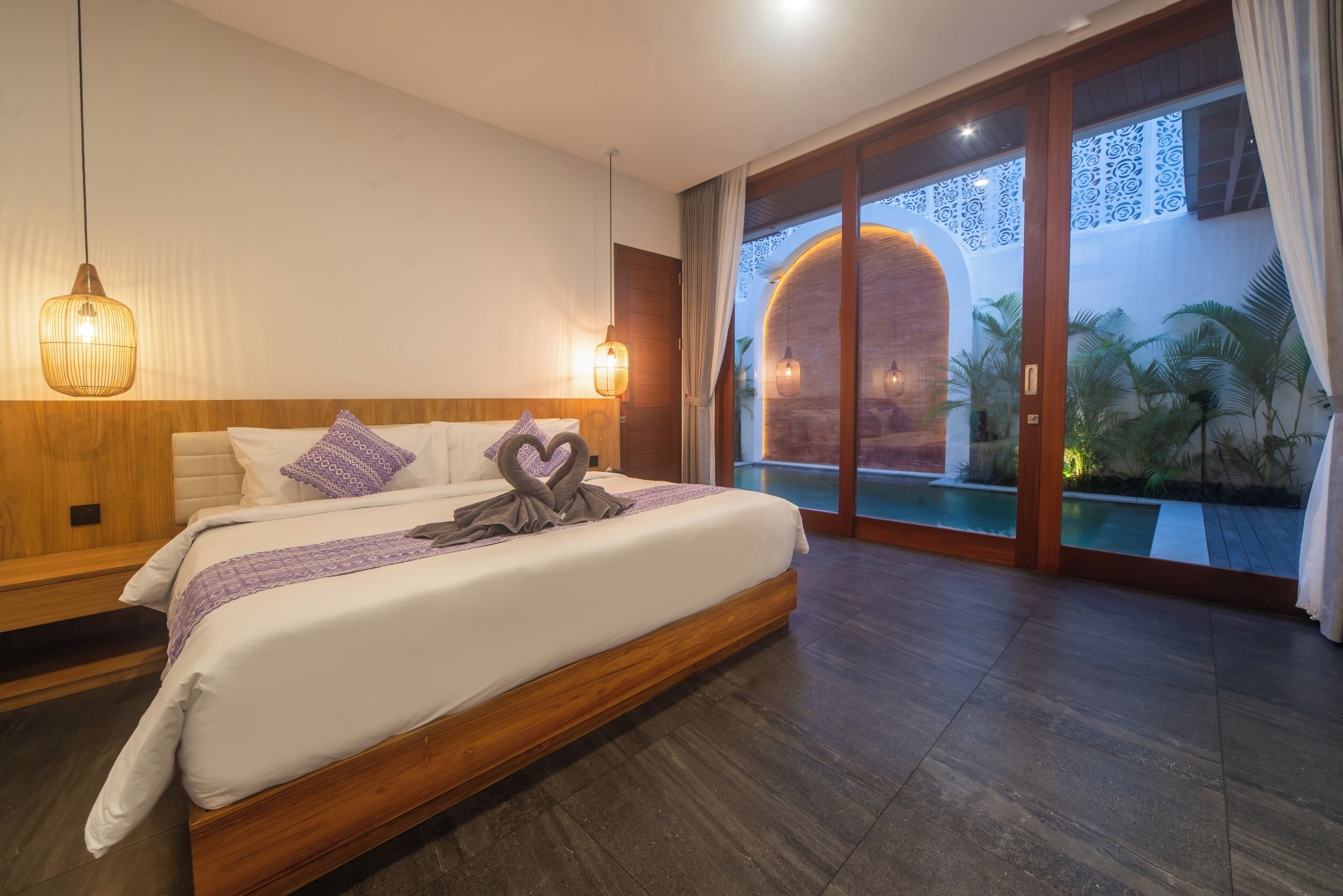 A modern hotel room with a large bed, wooden headboard, and a view of a swimming pool through glass doors.