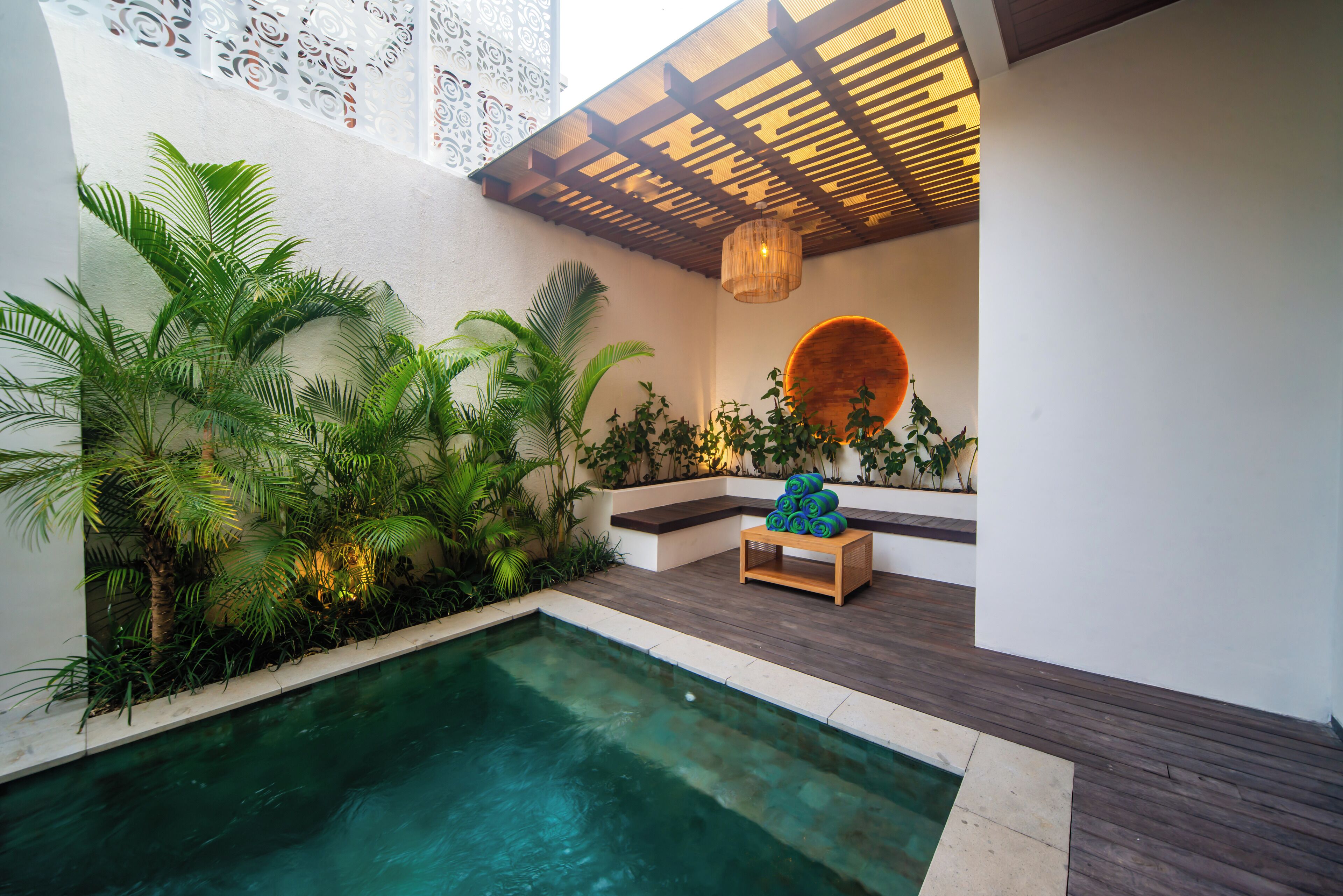 A pool area with a wooden deck, lush greenery, and a seating area with a round mirror and a hanging light fixture.