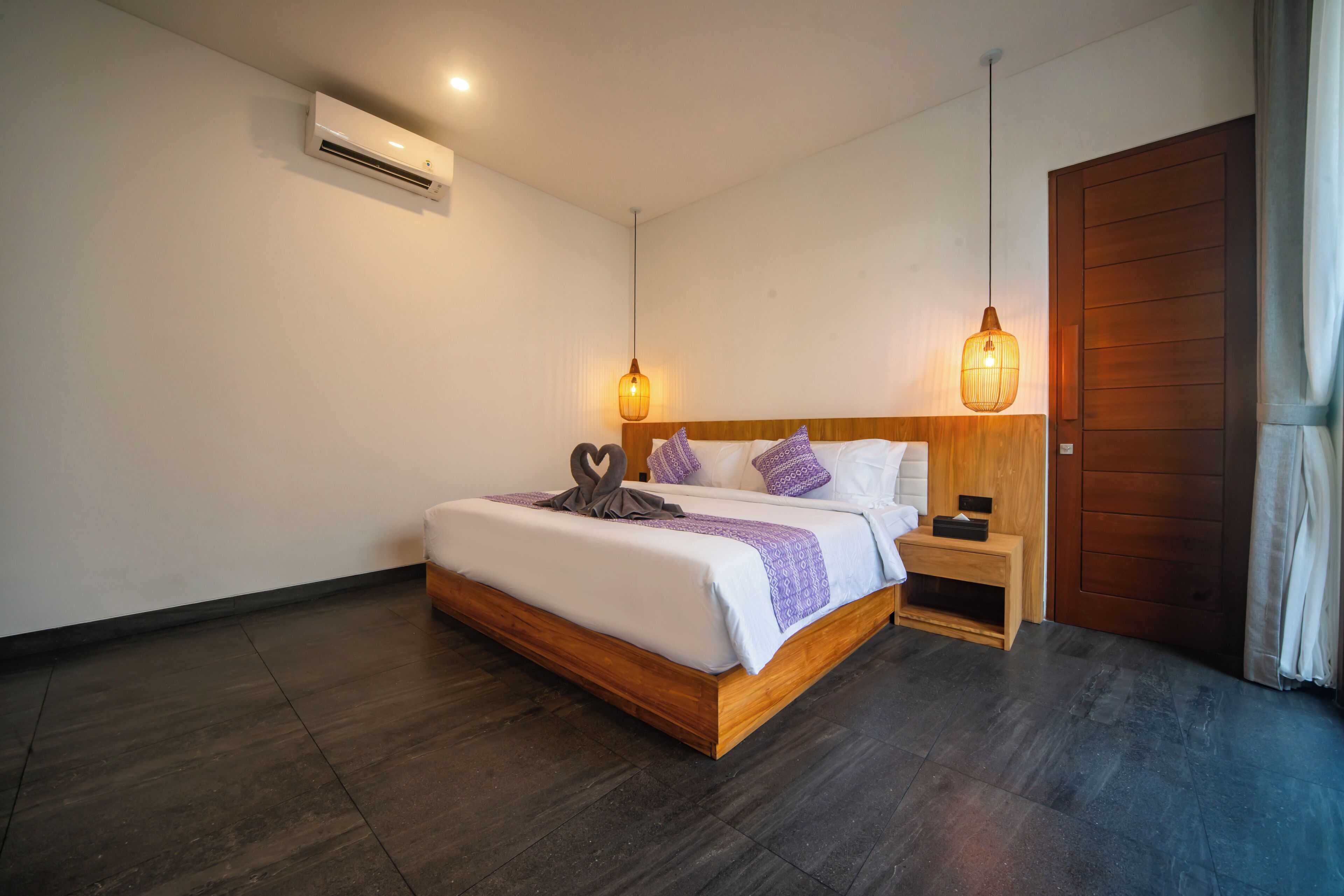 A modern hotel room with a wooden bed, a nightstand, an air conditioning unit, and a wooden door.