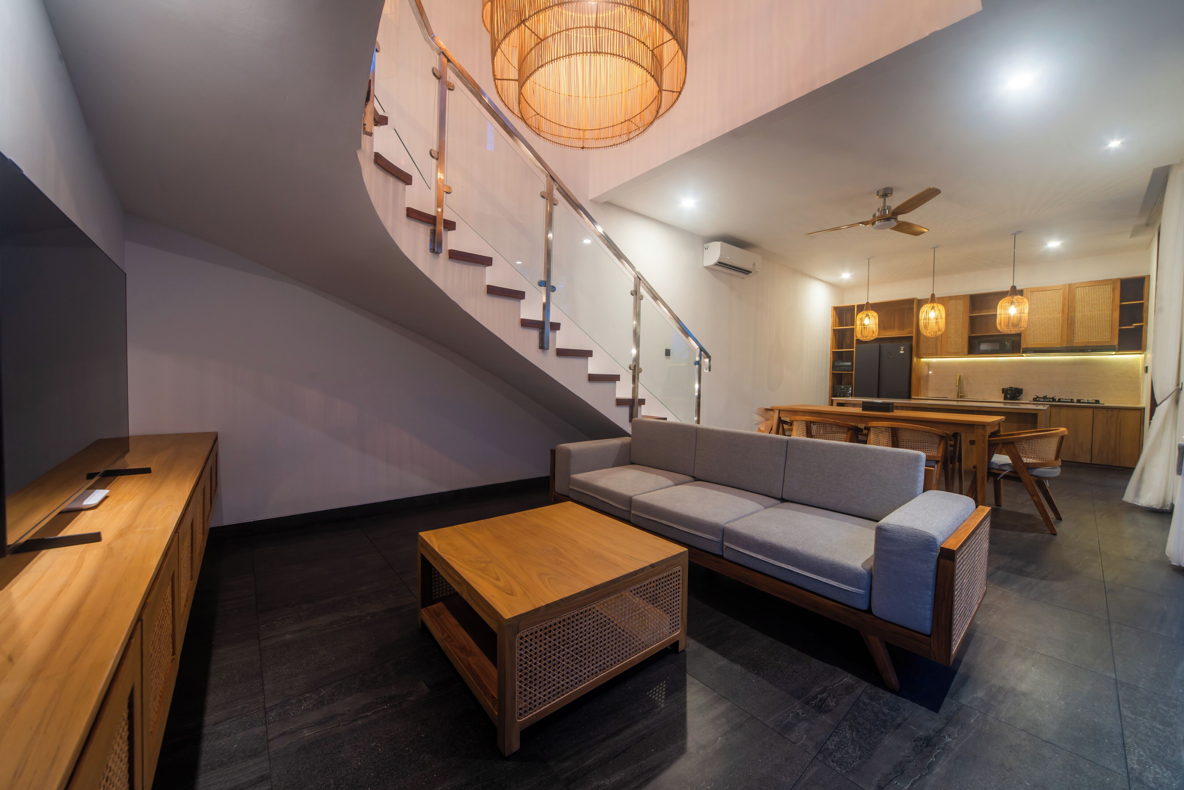 Un interior moderno con una escalera curva, una mesa de centro de madera, un sofá gris y una zona de cocina con armarios de madera y luces colgantes.