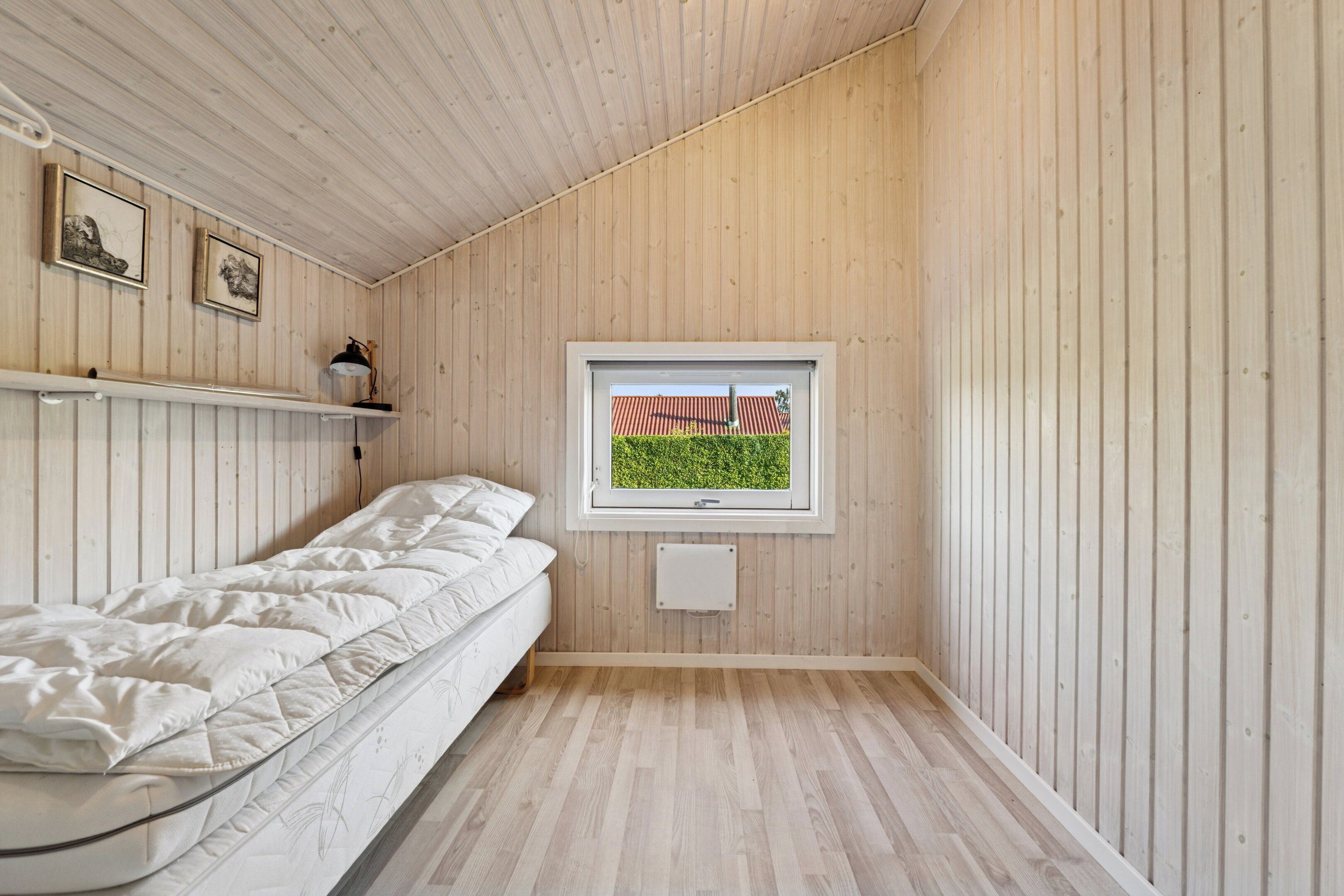 A room with a bed, a window with a view of a red-roofed building, wooden walls, and a shelf with a lamp.