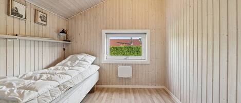 A room with a bed, a window with a view of a red-roofed building, wooden walls, and a shelf with a lamp.