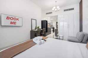 Quarto de hotel moderno com cama, escrivaninha, espelho, guarda-roupa e cozinha compacta.