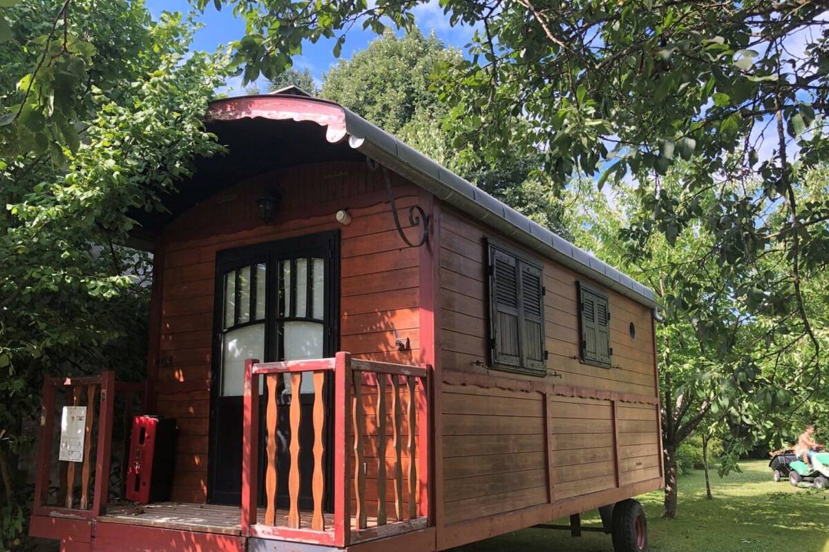 Un camping-car en bois, doté d’un porche rouge et de fenêtres à cadre noir, est garé dans une zone herbeuse bordée d’arbres.