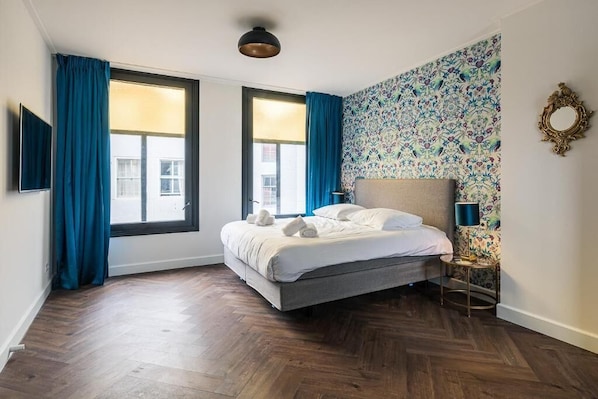 A bedroom with a bed, two windows with blue curtains, a patterned wall, and a small bedside table with a lamp. - De Bij Middelburg (Middelburg)
