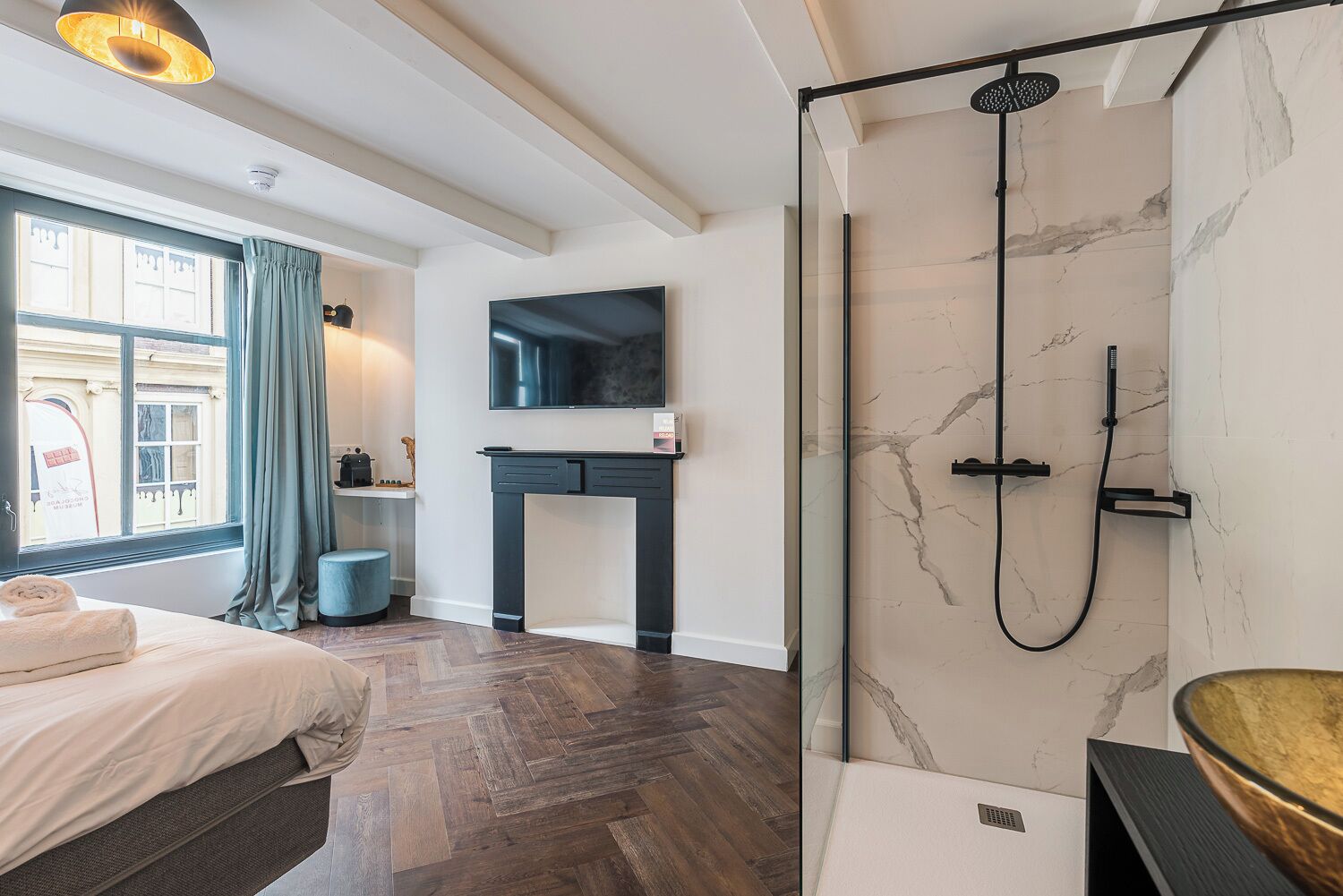 Et moderne hotelværelse med en gulv i fiskebensmønster af træ, et stort vindue med gardiner, en seng, et lille rundt bord, et sort konsolbord, et brusekabine i marmor og et ophængt fjernsyn.