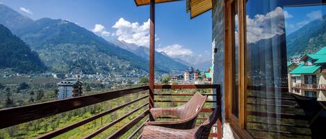 A balcony with wicker chairs, a wooden ceiling, and a view of a mountainous landscape with buildings in the distance.