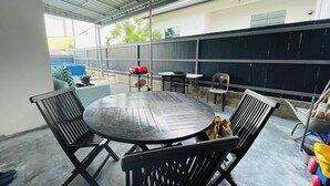Een overdekt terras met een ronde tafel en vier stoelen, een barbecue en een kleine eettafel met stoelen.
