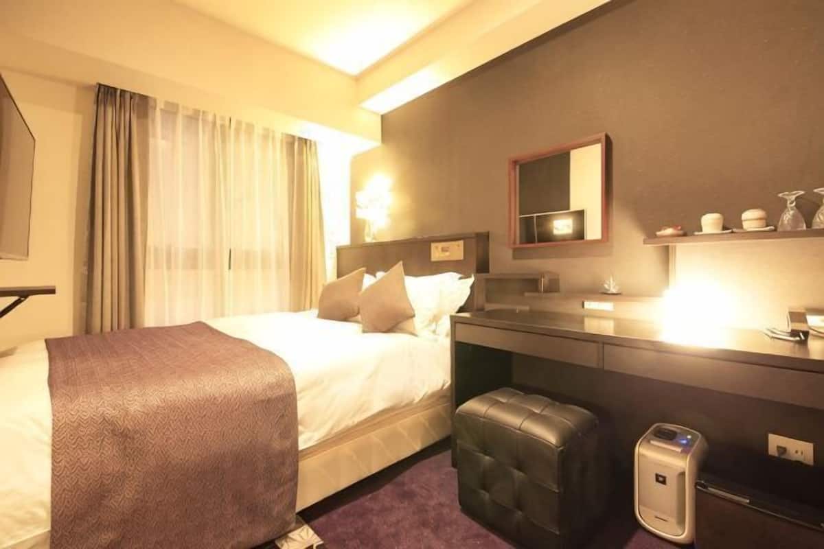 A hotel room with a bed, a desk, a mirror, a shelf with toiletries, and a suitcase.