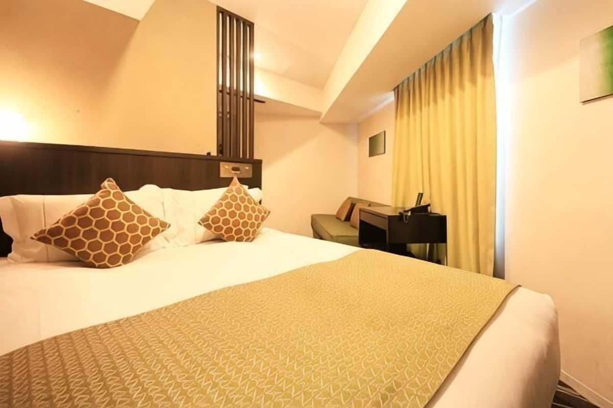 A hotel room with a bed, pillows, a bedside table, a lamp, and a window with curtains.