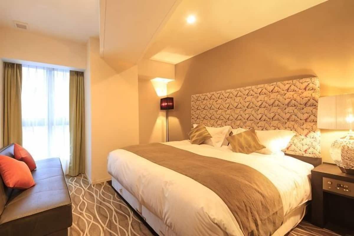 A hotel room with a large bed, a bedside table, a lamp, and a view of the outdoors.