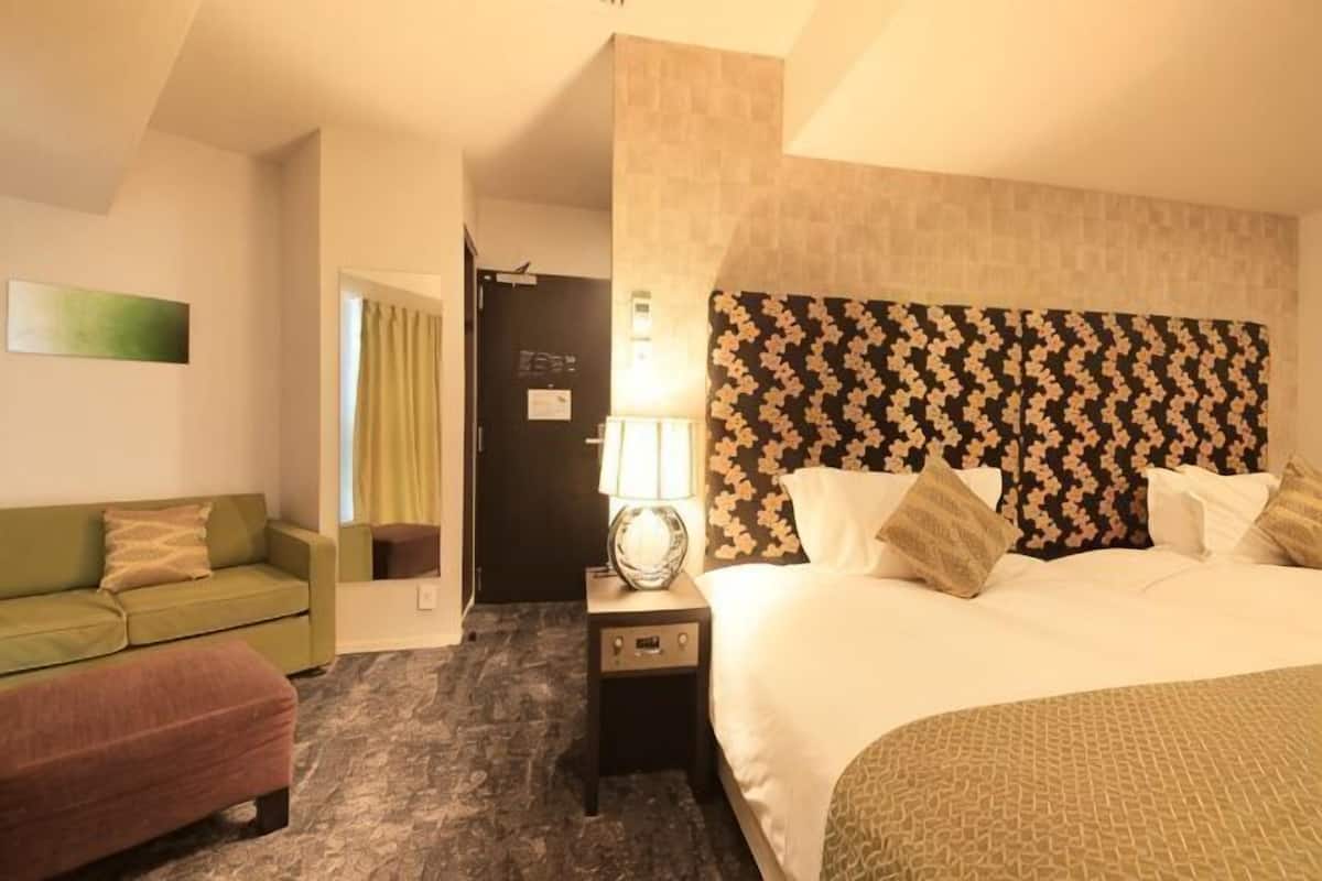 A hotel room with a bed, a nightstand with a lamp, a green sofa, and a red chair.