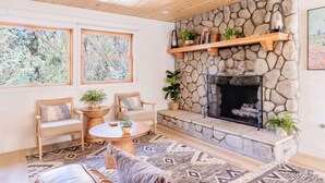 Un salon chaleureux avec une cheminée en pierre, des meubles en bois et de grandes fenêtres donnant sur la forêt.
