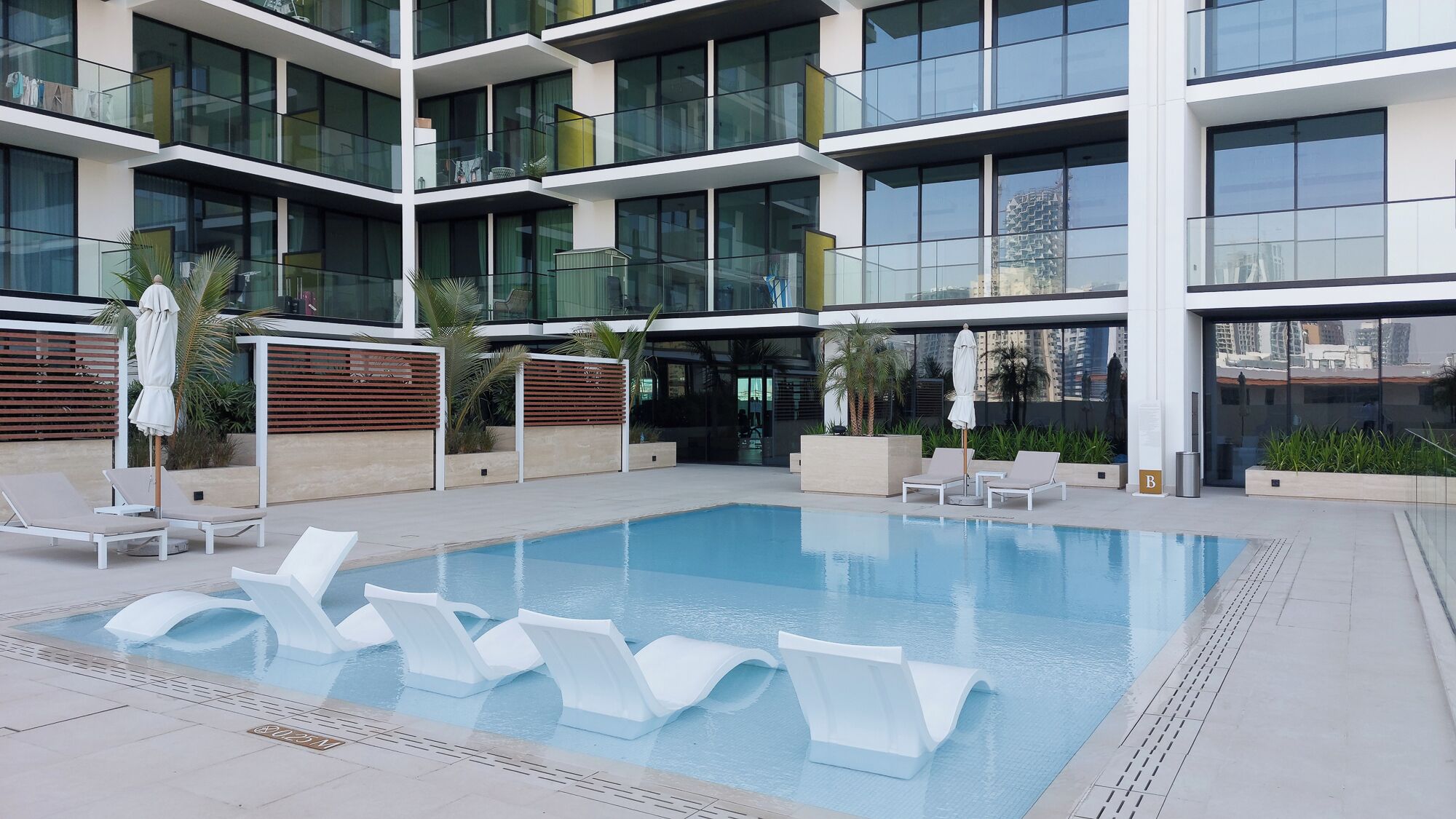 Un edificio moderno con piscina en la azotea, sillas de descanso y fachada de vidrio.