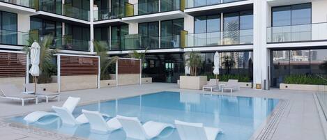 Un edificio moderno con piscina en la azotea, sillas de descanso y fachada de vidrio.