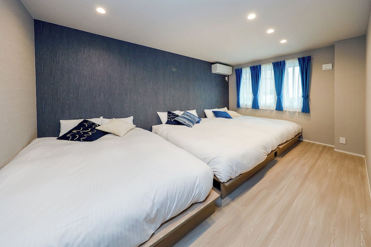 A hotel room with two beds, a dark wall, a window with blue curtains, and a wooden floor.