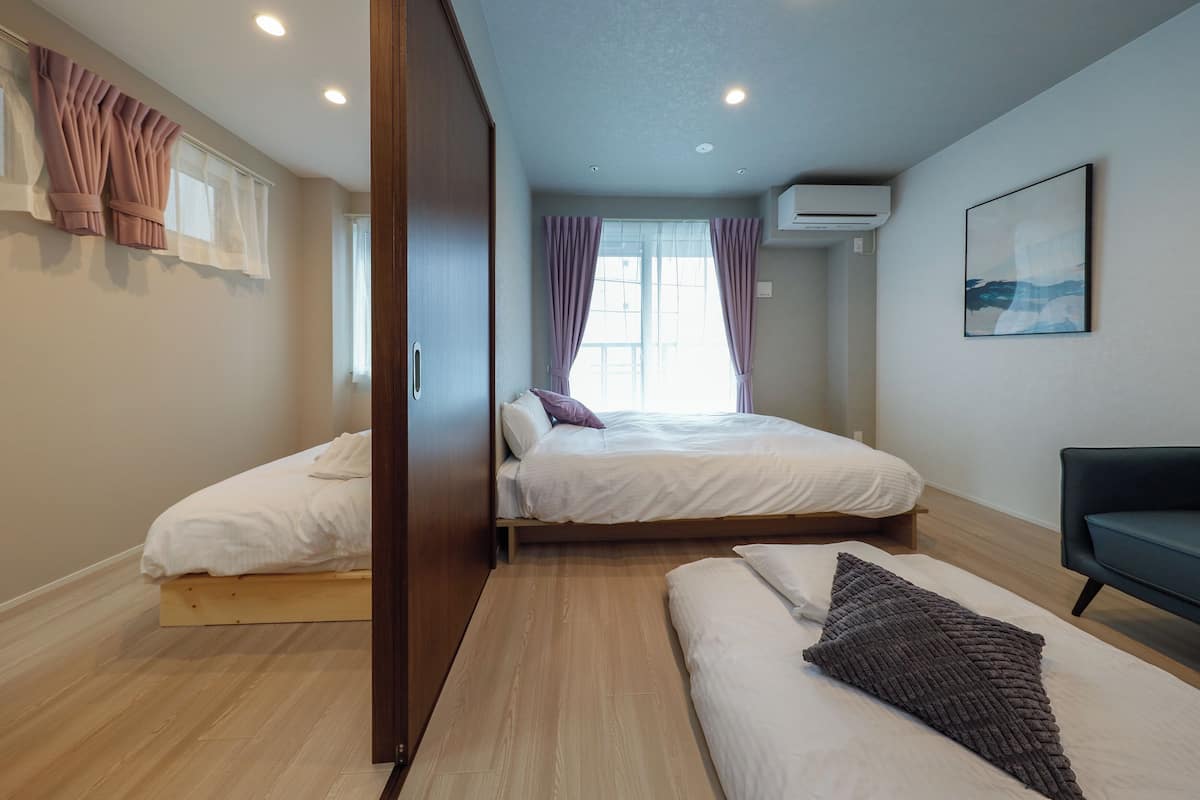 A bedroom with two beds, a sofa, a window with curtains, an air conditioning unit, and a painting on the wall.