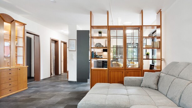 Un salon moderne avec un canapé d’angle, des étagères en bois et un coin cuisine à l’arrière-plan.