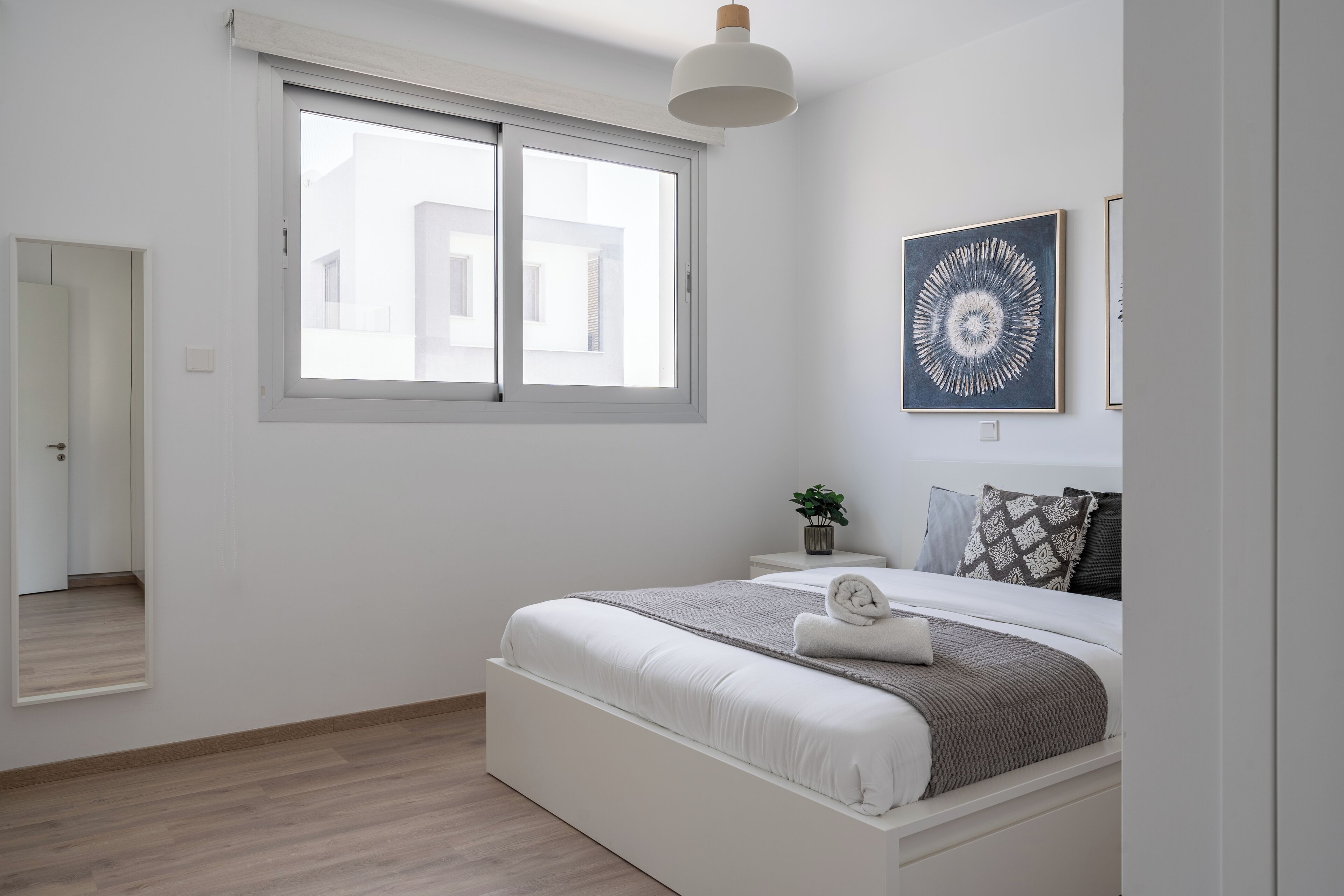 A bedroom with a large window, a bed with white bedding, a small bedside table with a plant, and a wall-mounted artwork.