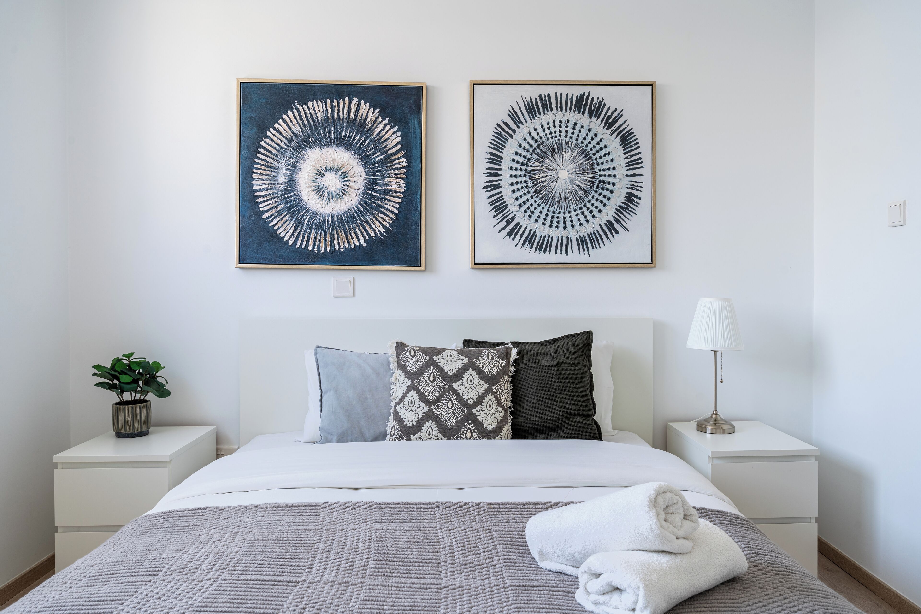 Une chambre à coucher bien rangée, avec un lit, des oreillers, une lampe de chevet et deux œuvres d’art encadrées accrochées au mur.