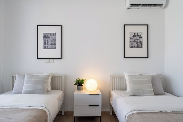 A bedroom with two beds, each with a white headboard, a bedside table with a lamp, and framed pictures on the wall.