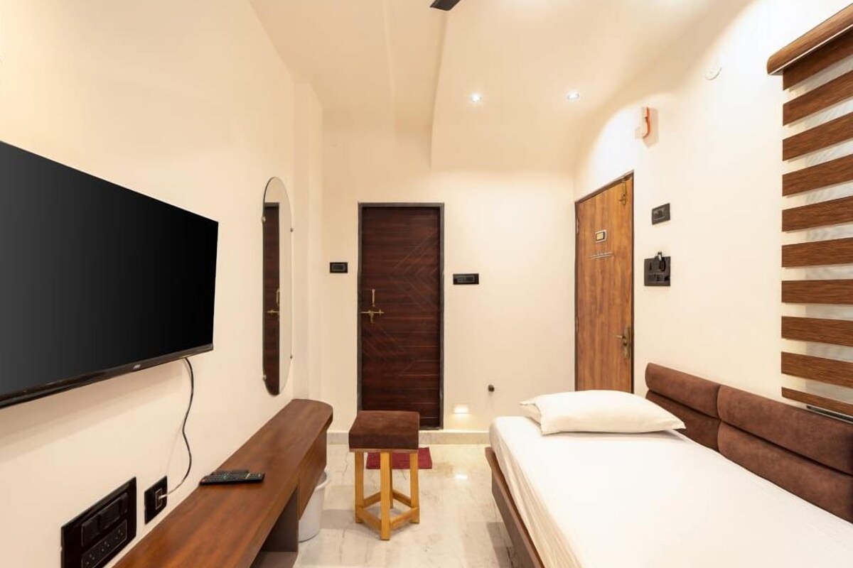 Een hotelkamer met een flatscreen televisie aan de muur, een houten bankje, een bed met een bruin hoofdeinde en een houten deur.