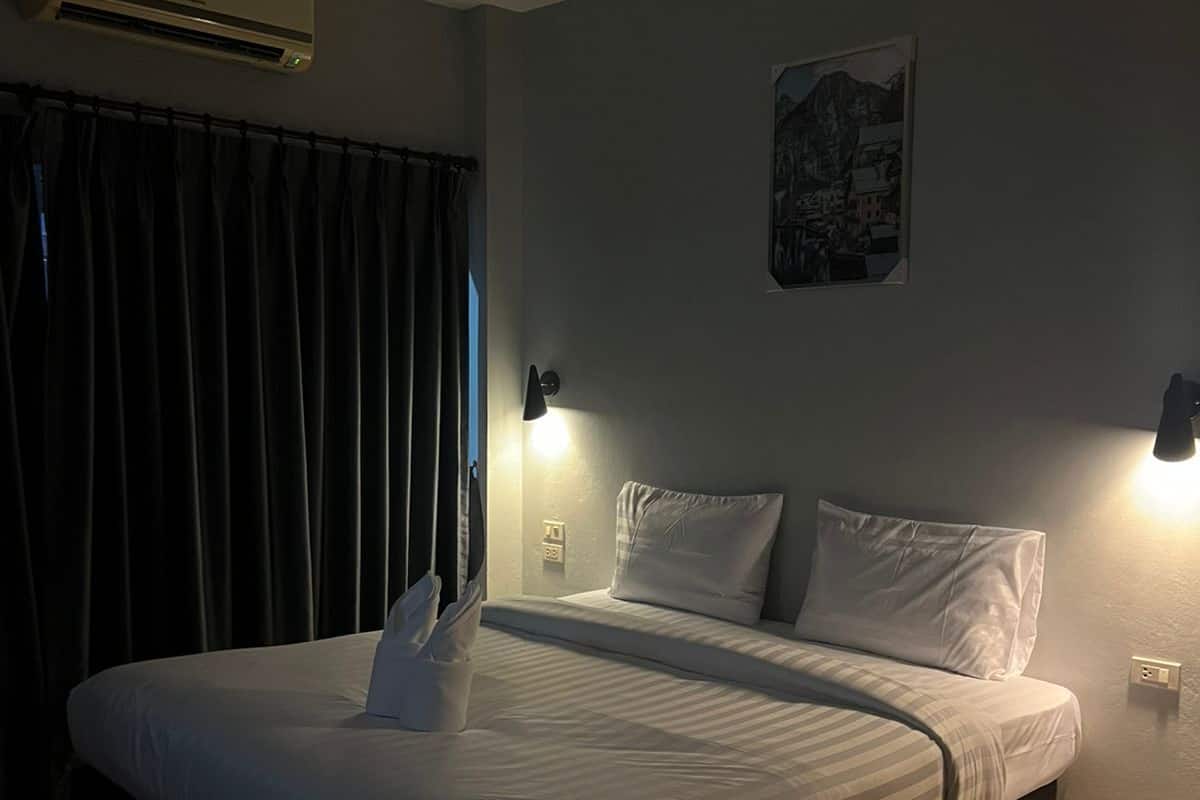 A hotel room with a bed, two bedside lamps, an air conditioner, and a painting on the wall.