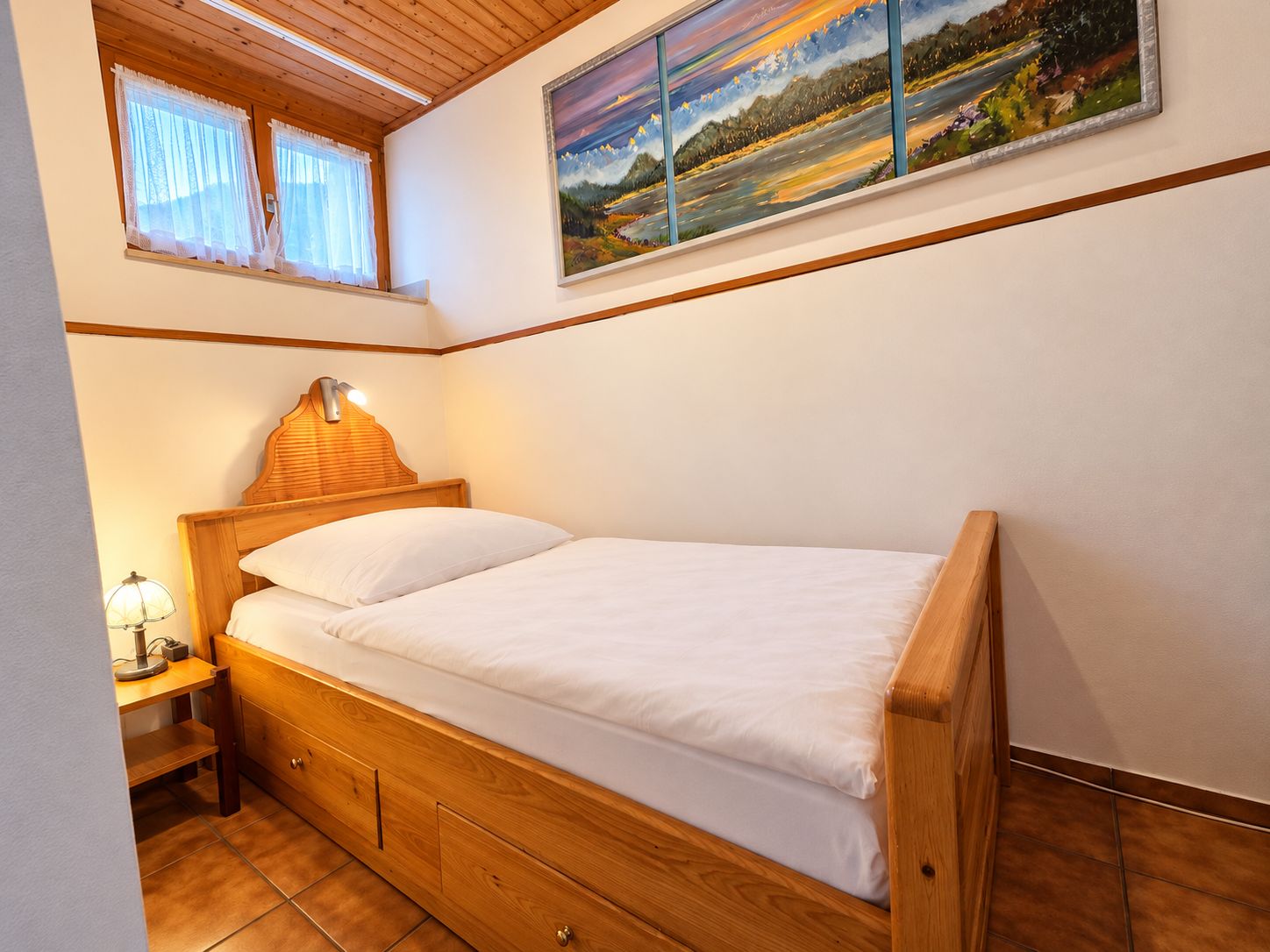 Een enkel bed met houten frame en hoofdeinde, een nachtkastje met een lamp, en een raam met gordijnen.