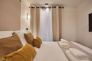 Quarto de hotel com cama bem arrumada, um conjunto de toalhas e vista pela janela com cortinas.