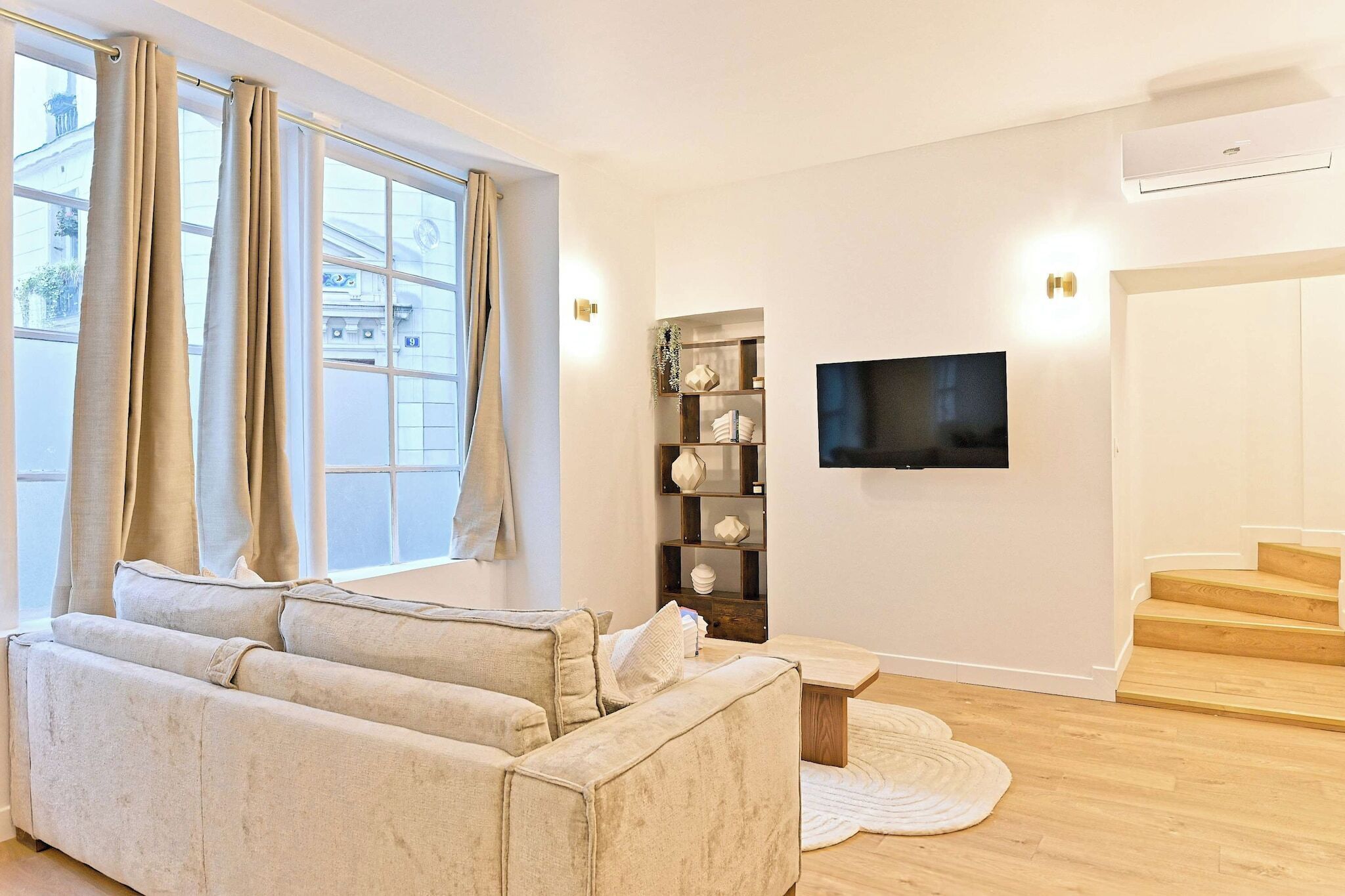 A living room with a beige sofa, a small coffee table, a TV mounted on the wall, and a staircase leading to an upper floor.