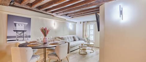 A modern living room with exposed wooden beams, a sofa, a dining table with chairs, and a wall-mounted light fixture.