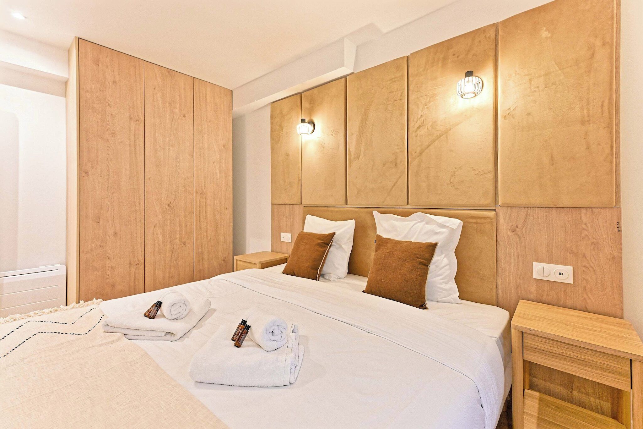 Un lit bien fait, avec du linge de lit blanc, deux oreillers marron et un plaid blanc. Il y a deux tables de chevet en bois, chacune surmontée d’une petite lampe. La tête de lit est également en bois et est dotée d’un éclairage intégré.