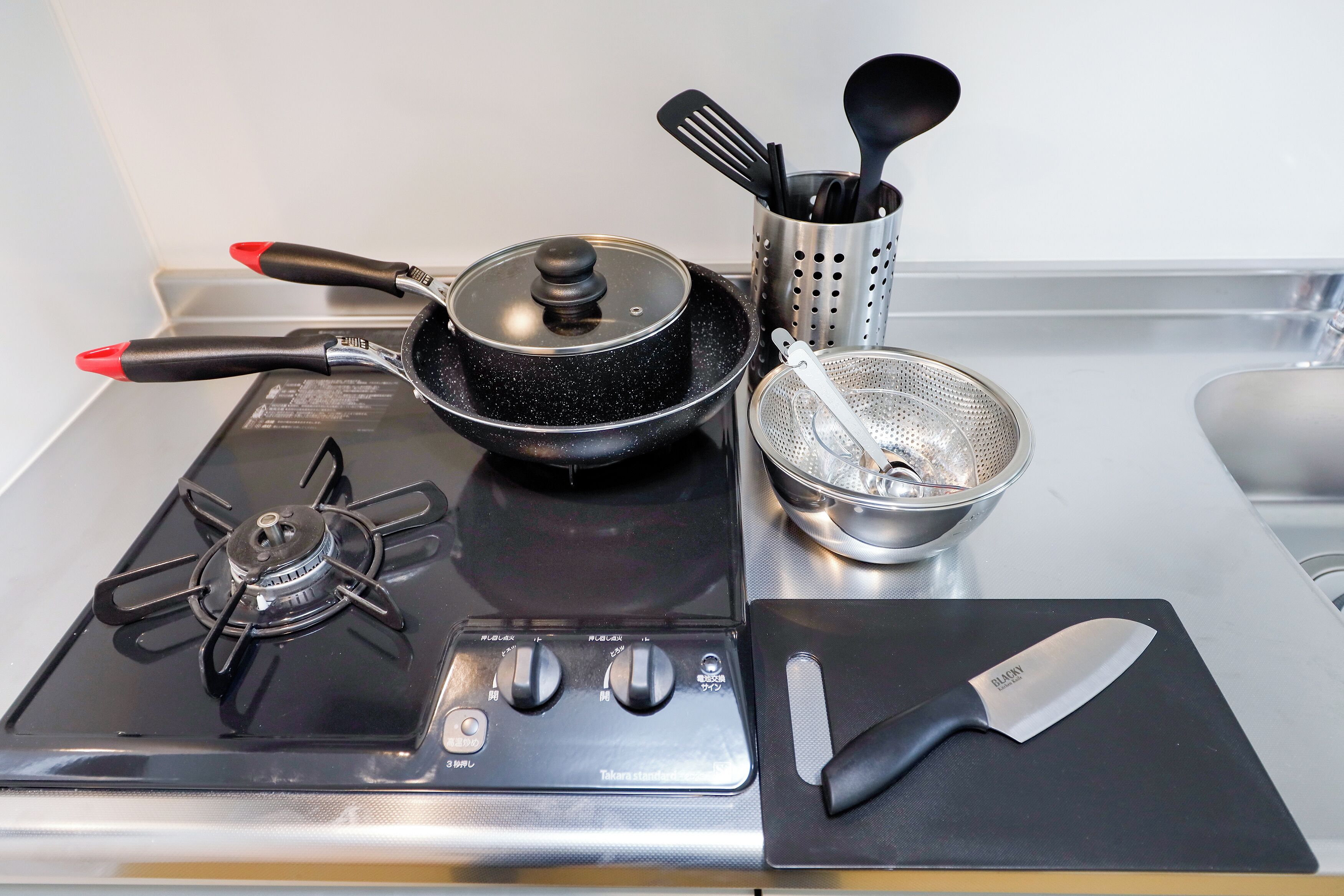 Una cocina con estufa a gas, una sartén, un colador, un soporte para espátulas y un cuchillo sobre una tabla de cortar.