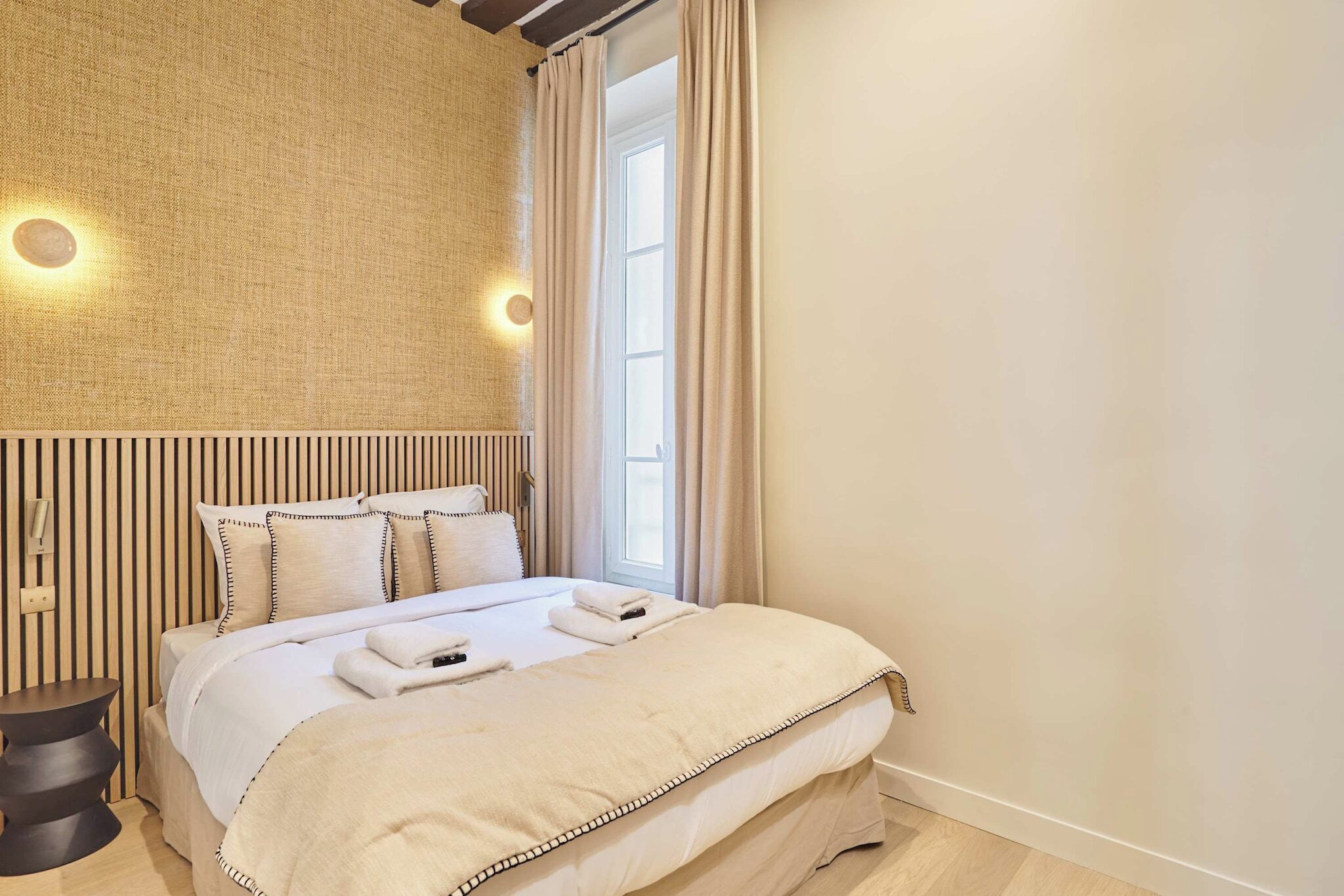 A hotel room with a large bed, a wall-mounted light, a window with curtains, and a textured wall.