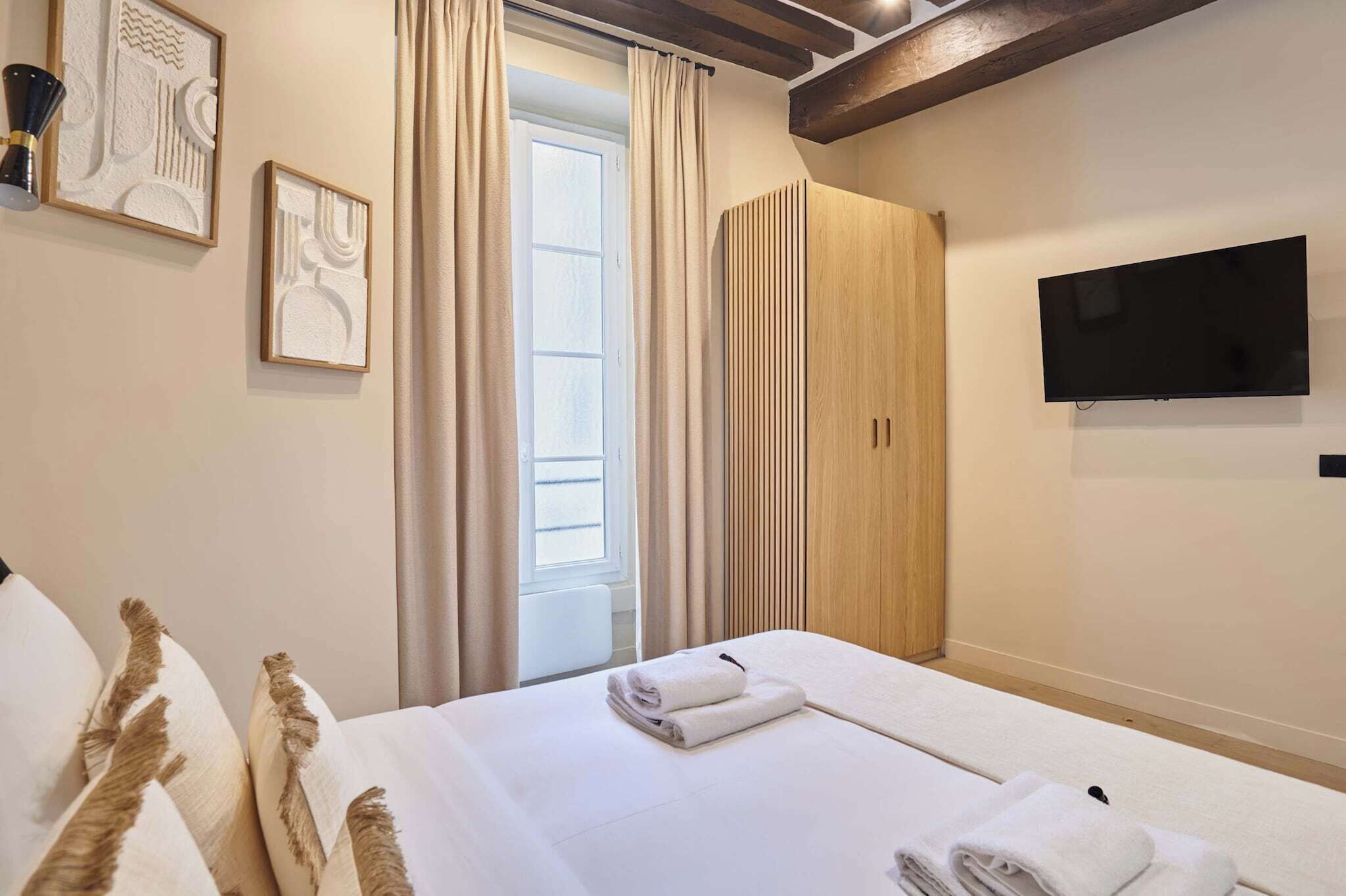 A hotel room with a bed, a TV mounted on the wall, a wardrobe, and a window with curtains.