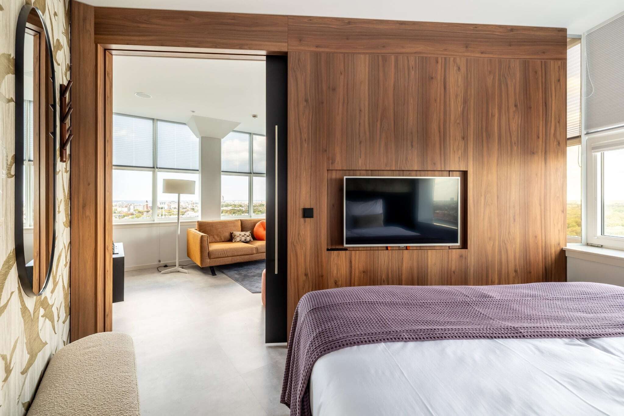 Een moderne slaapkamer met een groot bed, een televisie bevestigd aan een houten wand en uitzicht op de stad door grote ramen.