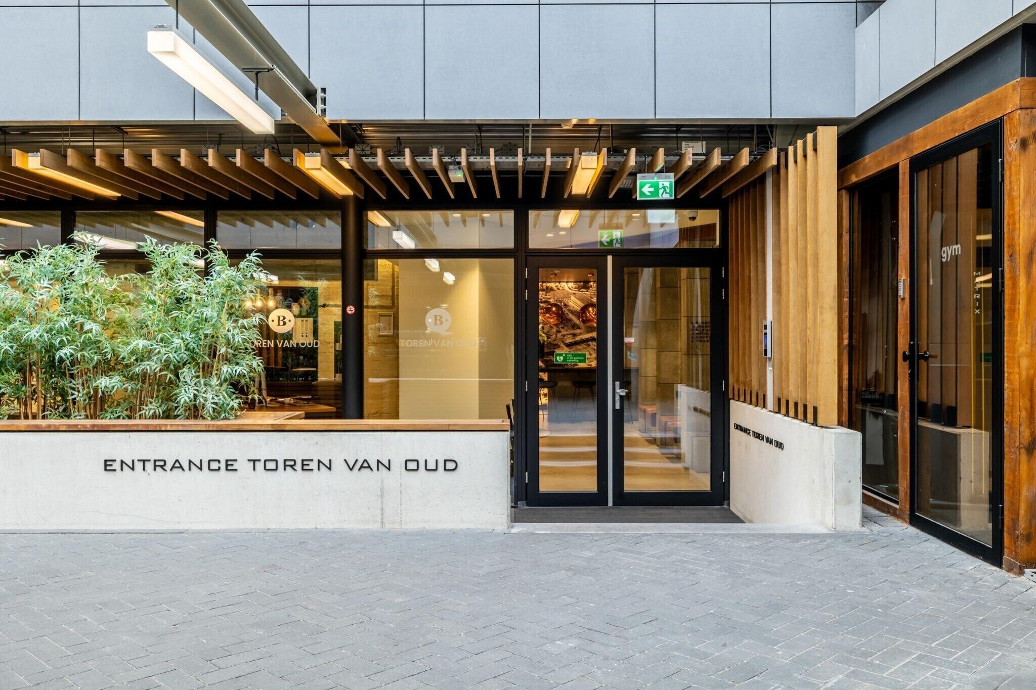De ingang van een gebouw met een modern ontwerp, met een glazen deur en een houten bank ervoor.