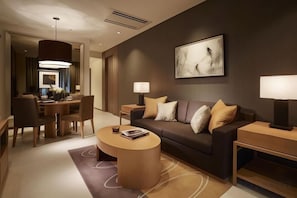 Una habitación de hotel moderna con un sofá de color oscuro, una mesa redonda, una mesita de noche con una lámpara y un comedor con candelabro.