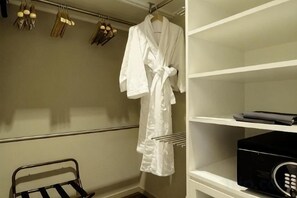 Hotelkamer met een witte badjas aan een rek, een zwarte afstandsbediening op een plank, en een metalen rek met handdoeken.