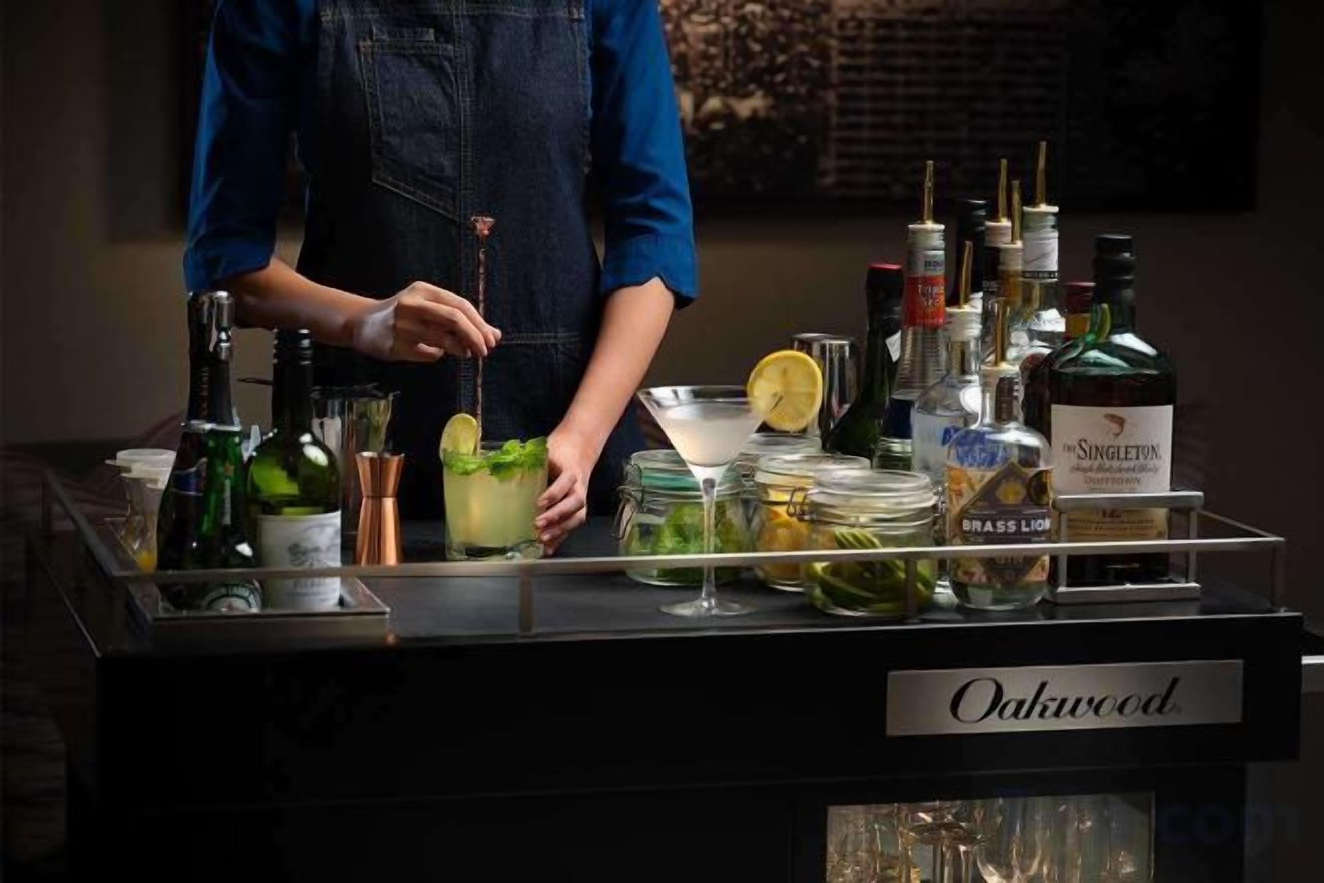 Zona de bar con un bartender preparando una bebida, una variedad de botellas y un letrero que dice 'Oakwood'.