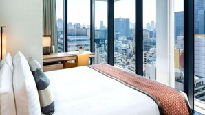 Habitación de hotel con una cama grande, un escritorio y vistas de la ciudad a través de amplios ventanales.