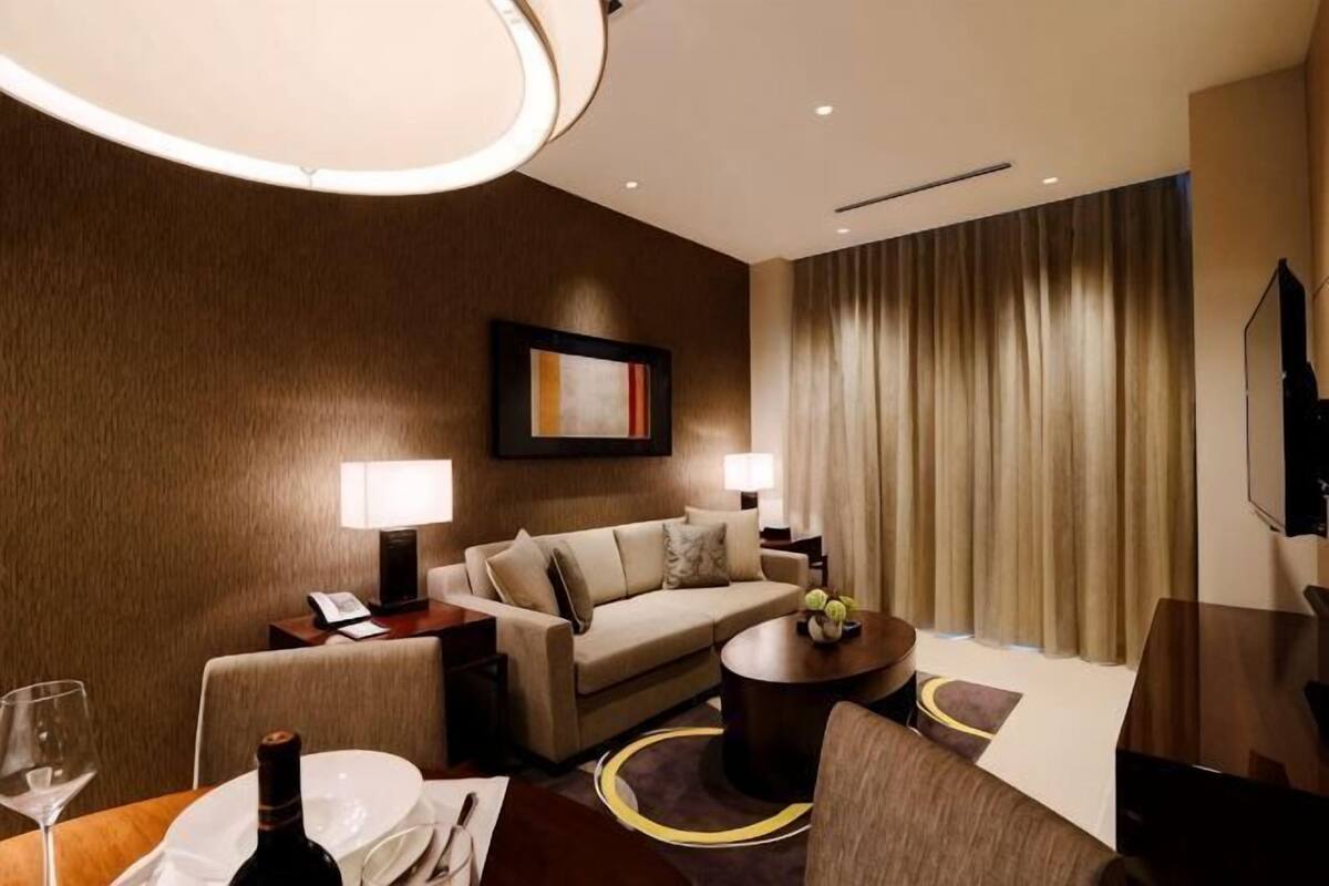 Une chambre d’hôtel avec un canapé, une table basse, une table à manger avec une bouteille et des verres, une télévision et une grande fenêtre avec des rideaux.