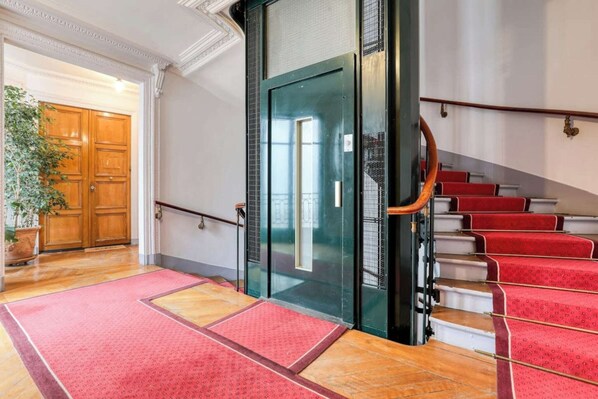 A hotel lobby with a wooden door, a glass elevator, and red carpeted stairs.