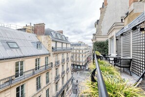 A narrow street with buildings on both sides, featuring balconies and a small outdoor seating area with tables and chairs.