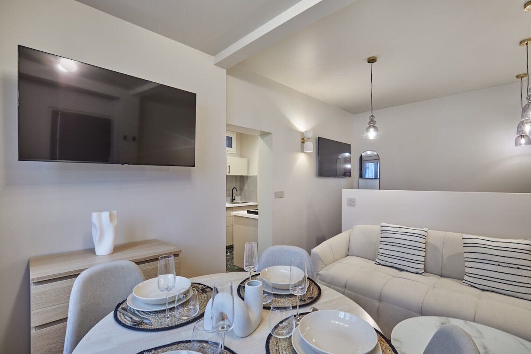 A modern living room with a sofa, dining table set with plates and glasses, a flat-screen TV mounted on the wall, and pendant lighting.